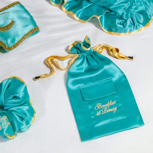 The Breakfast at Drowsy Turquoise Carry Pouch by drowsysleepco, featuring gold embroidery and a gold ribbon, sits on a white surface among matching teal satin accessories with gold trim like a sleep mask and scrunchie.