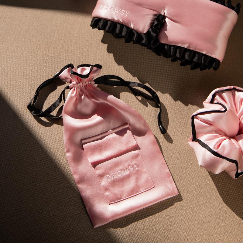 A pink drowsysleepco Sleep and the City Silk Carry Pouch with a front pocket, shown with a matching eye mask and black-trimmed scrunchie, all arranged on a beige surface.