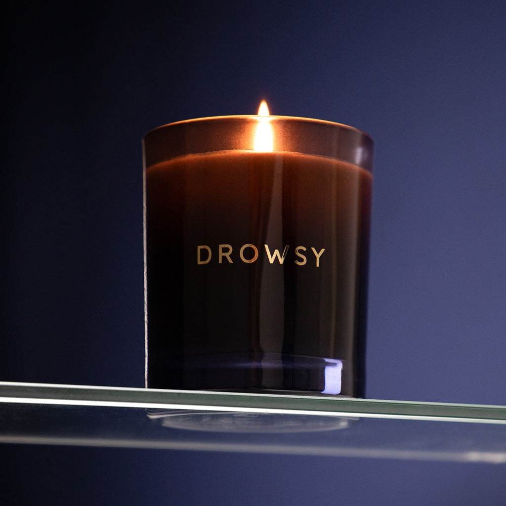 The Sleep Ritual Candle 220g, in a dark brown glass holder labeled DROWSY, rests on a glass shelf against a deep blue backdrop—perfect for your bedtime ritual.