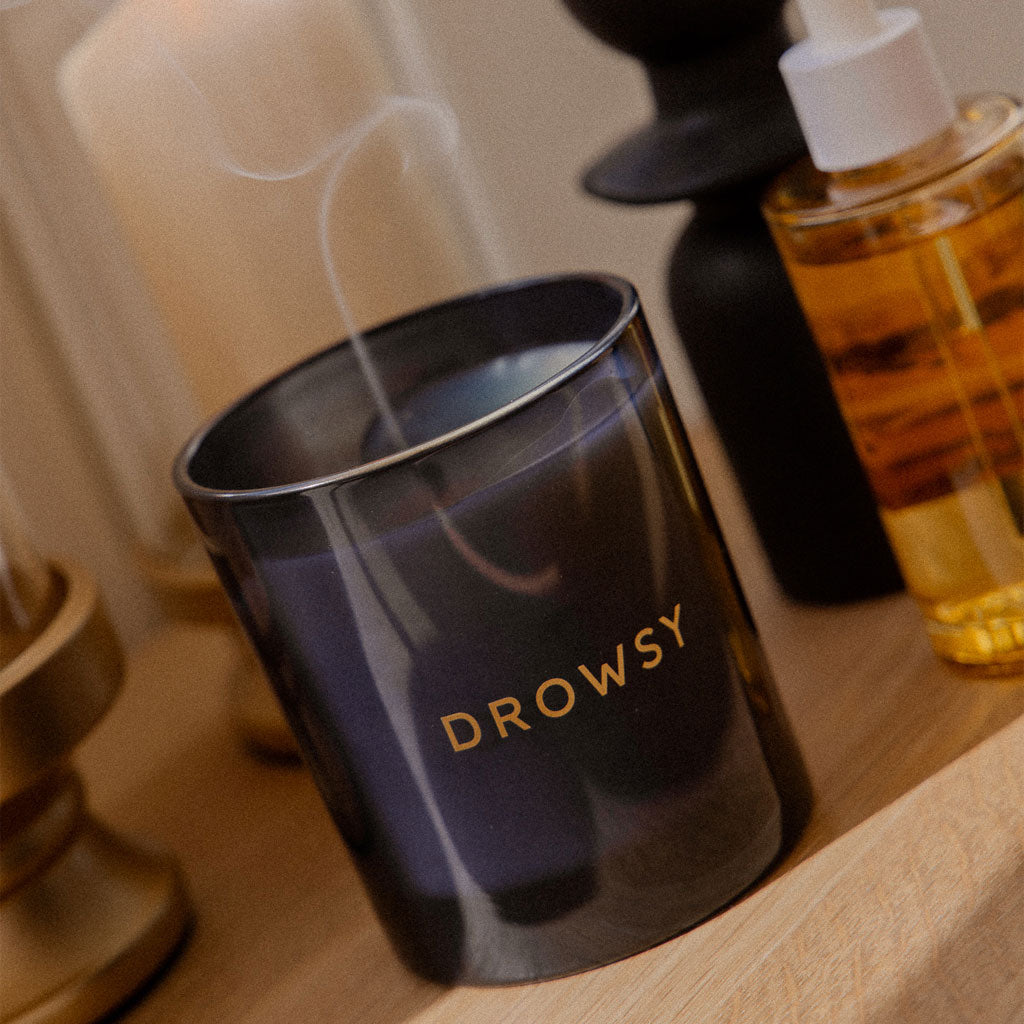 A black Sleep Ritual Candle 220g jar with DROWSY in gold lettering sits on wood, releasing a thin smoke trail—ideal for bedtime. Nearby are a gold holder, a larger candle, and a yellow bottle with a white cap.
