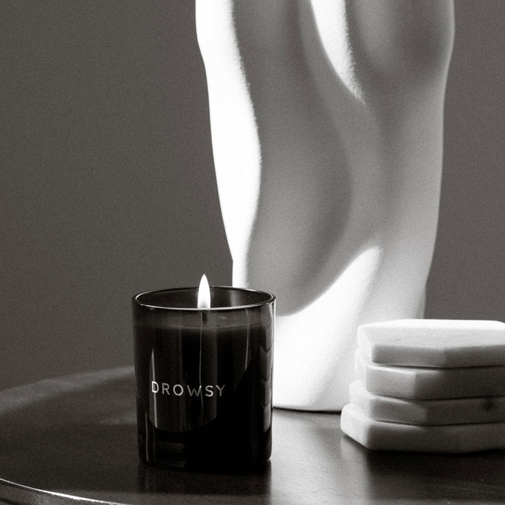 The Sleep Ritual Candle 220g in black burns on a round table beside three stacked white coasters and a white abstract sculpture, all aglow in soft, moody lighting—an ideal accent for your bedtime ritual.