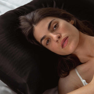 A woman with long brown hair lies on a Black Jade Silk Pillowcase, looking at the camera in a white strap top—a luxurious essential for skincare and hair protection.
