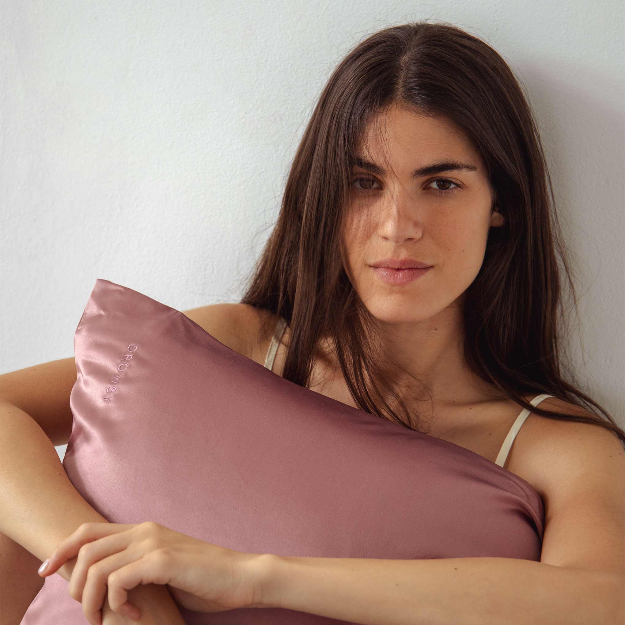 A woman with long brown hair sits against a white wall, calmly holding the drowsysleepco Sensual sleep bundle - Damask Rose, adding an extra touch of elegance as she looks at the camera.