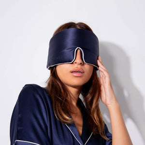 A woman wearing Midnight Blue - Pipe Dreams Edition satin pajamas holds the matching adjustable sleep mask over her eyes against a plain white background.