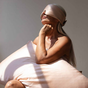 An older woman with long gray hair sits serenely in sunlight, wearing a satin sleep mask and holding a Sunset Pink Silk Pillowcase designed for hair protection and skincare.