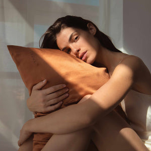 A woman in a light top hugs the Pumpkin Spice Silk Pillowcase while sitting in soft natural light, shadows on her face and body. She looks relaxed, enjoying this skincare and beauty essential.