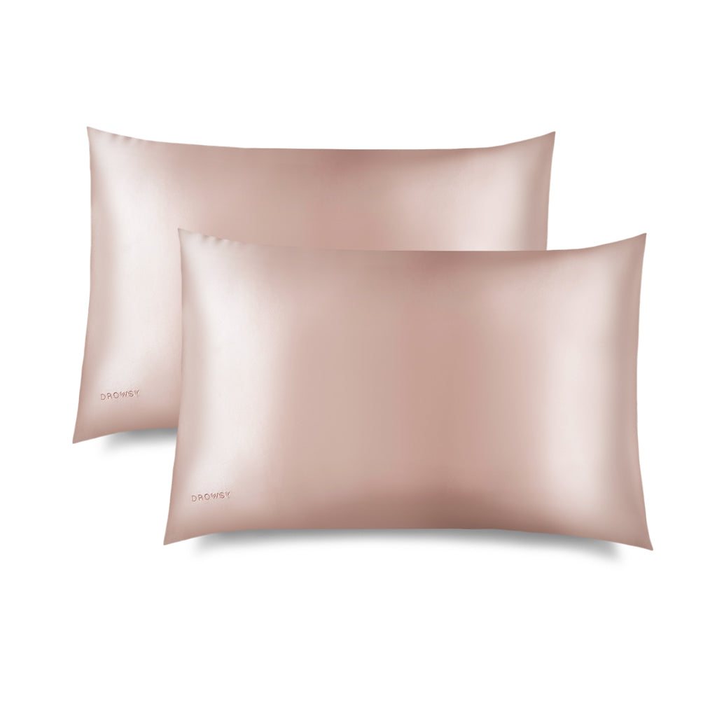 Two Sunset Pink Silk Pillowcases with a smooth, shiny finish are stacked and slightly overlapping on a white background. Each hair-protecting pillowcase has the word "DROWSY" subtly embroidered in the corner.
