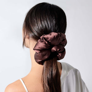 A woman with long, dark hair wears the drowsysleepco Cocoa Glow - Padded Scrunchie in a low ponytail. Dressed in a white, thin-strapped top, the oversized accessory pops against the simple, light background.