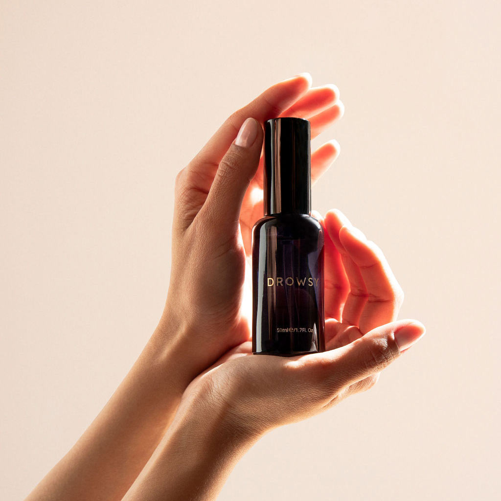 Two hands gently cradle a dark glass bottle labeled DROWSY against a soft, neutral background, with warm light highlighting the product.