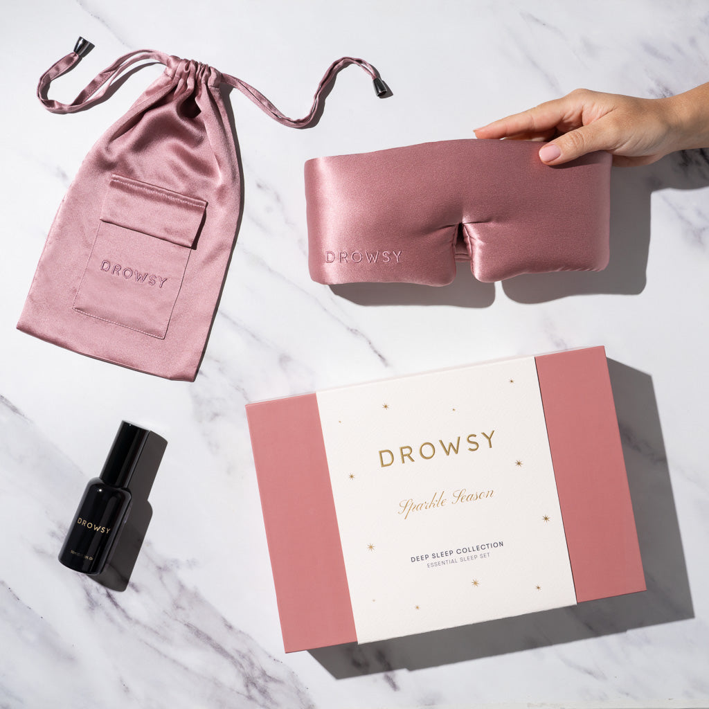 A hand holds the Damask Rose - Deep Sleep Collection silk sleep mask beside a pink silk pouch, a black deep sleep pillow spray bottle, and a pink and white gift box on a white marble surface.