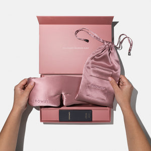 A person holds the Damask Rose - Deep Sleep Collection luxury silk sleep mask with matching pink carry pouch above an open pink gift box with “You deserve glorious sleep” inside the lid and a small product box nestled within.
