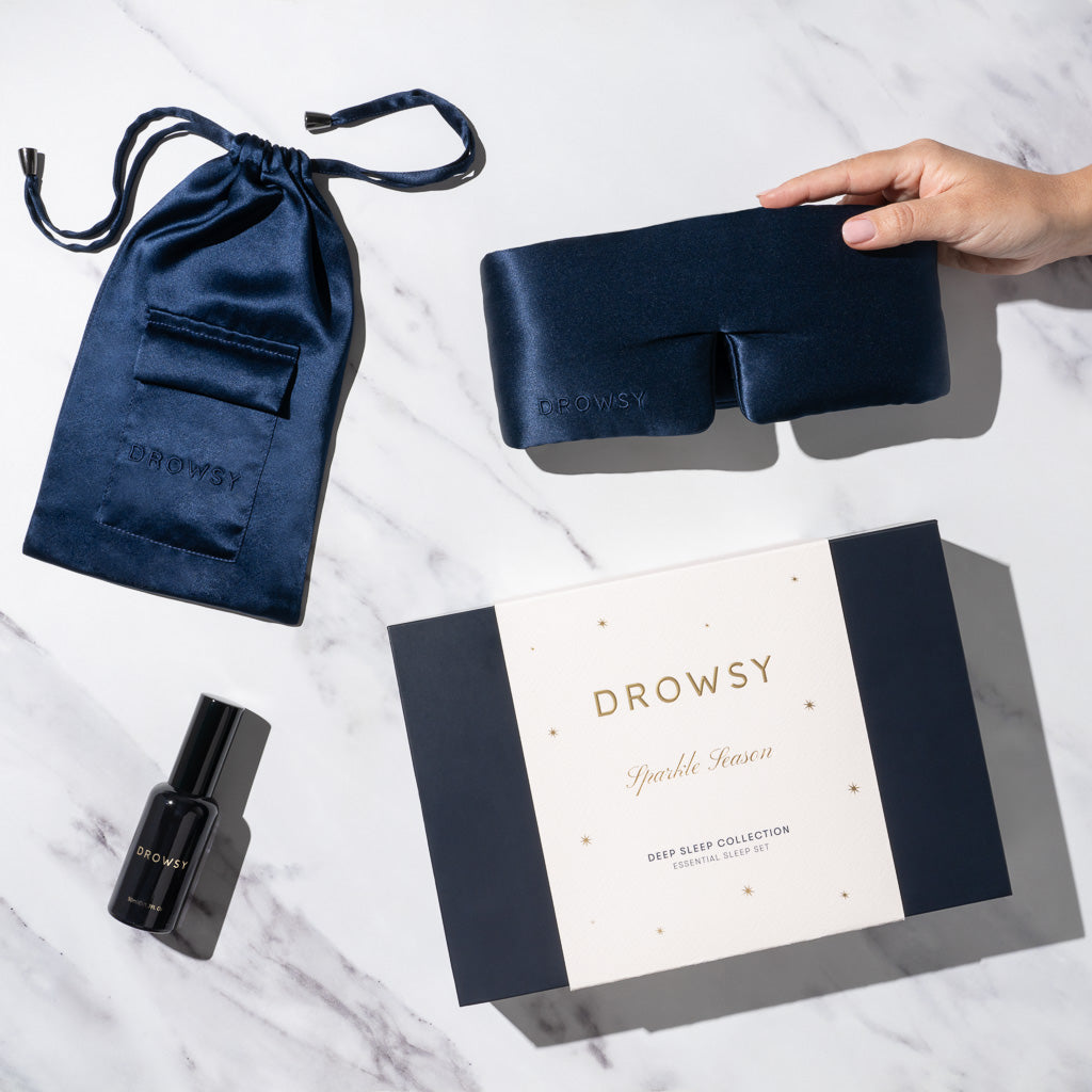 A hand holds a navy blue silk sleep mask above its pouch, a deep sleep pillow spray, and a gift box labeled “Midnight Blue - Deep Sleep Collection,” displaying luxury sleep essentials on a marble surface.