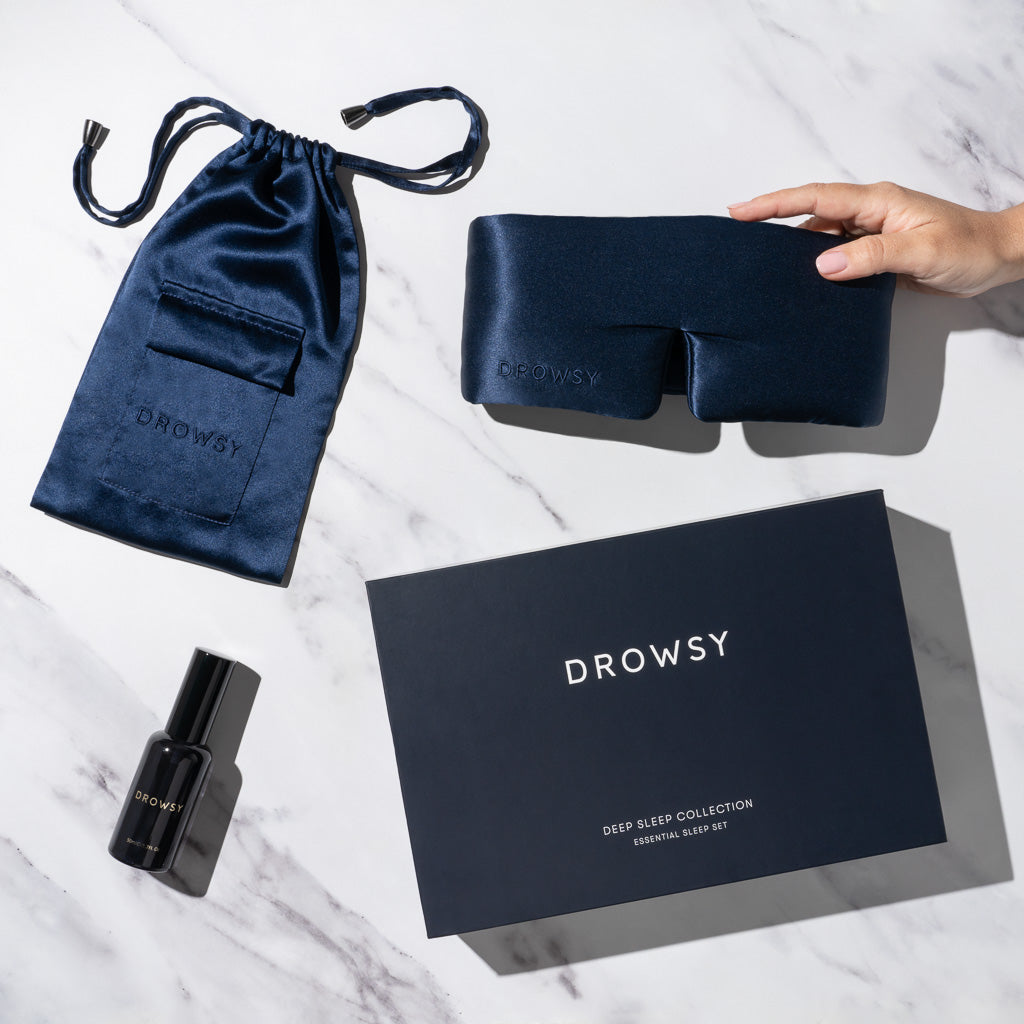 The Midnight Blue - Deep Sleep Collection, featuring a luxury silk sleep mask, matching pouch, pillow spray, and Drowsy-branded box, is arranged on white marble; a hand holds the mask—perfect for restful nights.