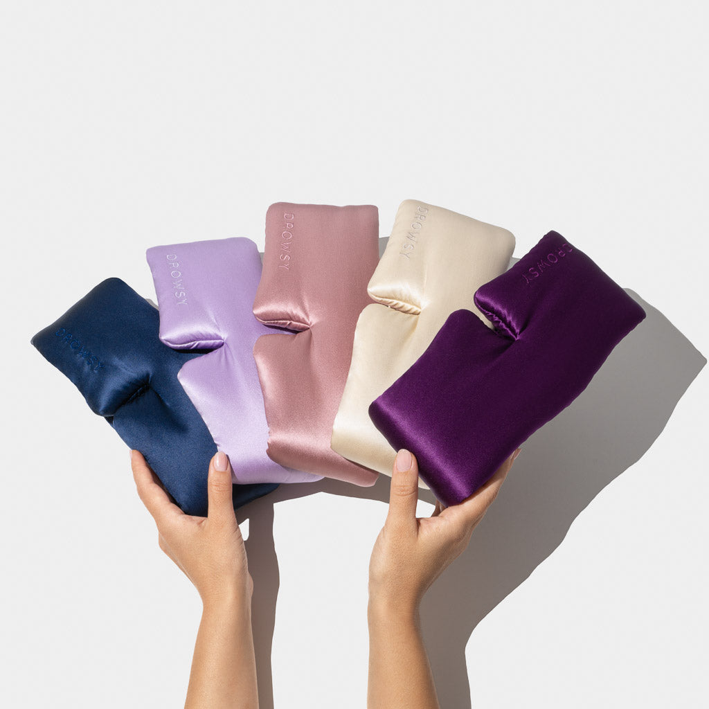 Two hands hold the Family Pack—five silky, rectangular eye masks in navy blue, light purple, rose pink, cream, and deep purple—fanned out against a plain white background.