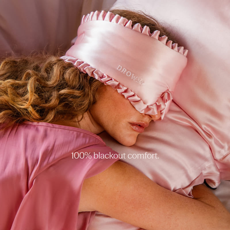 A woman with wavy hair lies on a bed wearing drowsysleepco’s Sleep and the City Powder Pink 100% mulberry silk sleep mask, matching pajamas, and resting on a pink silk pillowcase. Text reads "100% blackout comfort.