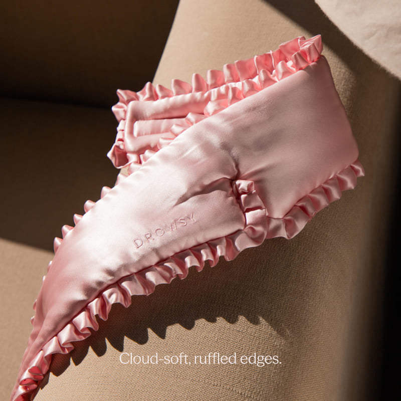 A light pink "Sleep and the City Powder Pink" mulberry silk sleep mask by drowsysleepco, with ruffled edges, rests on a beige cushion. "DROPSY" is embroidered on the mask. Below: Cloud-soft, ruffled edges.