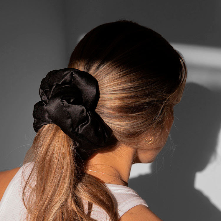 A woman with light brown hair wears a white sleeveless top and the drowsysleepco Silky skin & hair bundle - Black Jade scrunchie in her low ponytail, standing in soft light with her back to the camera.