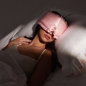 A woman sleeps peacefully in drowsysleepco's Manhattan Experience Bundle—a pink ruffled sleep mask and white embroidered pillow—soft light illuminating her as she rests in a white tank top.