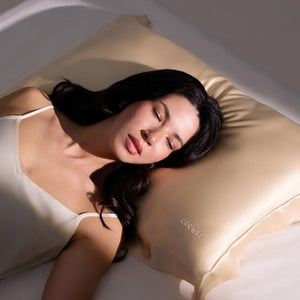A woman with long dark hair sleeps peacefully on a bed, resting her head on the us.drowsysleepco Hyalo Pillowcase in Champagne Gold. She wears a cream-colored camisole and appears calm and relaxed.
