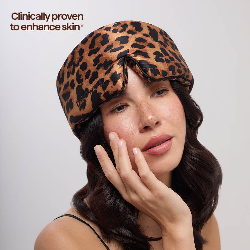 A woman with long wavy hair wears the Hotel Paradiso Wild Thing Sleep Mask by us.drowsysleepco, featuring a leopard print. Text in the top left corner reads, 