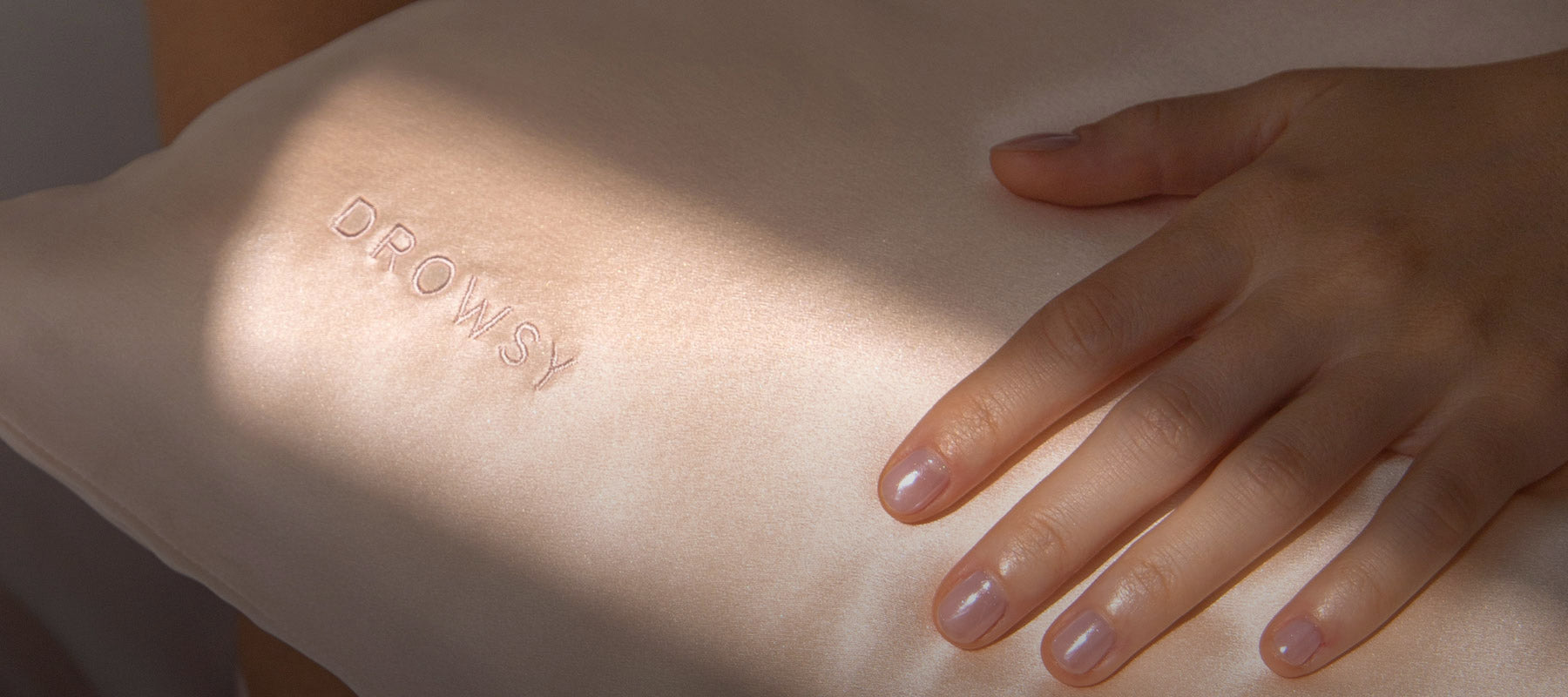 A hand with neatly manicured nails rests on a light pink silk pillow embroidered with the word DROWSY. Sunlight creates soft shadows across the fabric and hand.
