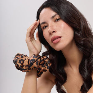 A woman with long, wavy dark hair rests her right hand on her face, wearing the Hotel Paradiso Wild Thing Padded Scrunchie—a leopard print silk accessory designed to promote hair thickness—against a light background, radiating calm confidence.