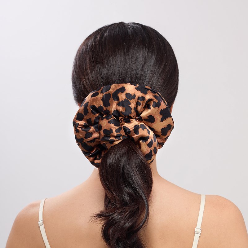 A woman with long, dark hair wears the Hotel Paradiso - Wild Thing Padded Scrunchie in leopard print on her low ponytail. She’s in a thin-strapped, light top against a plain background—ideal for promoting fuller-looking hair.