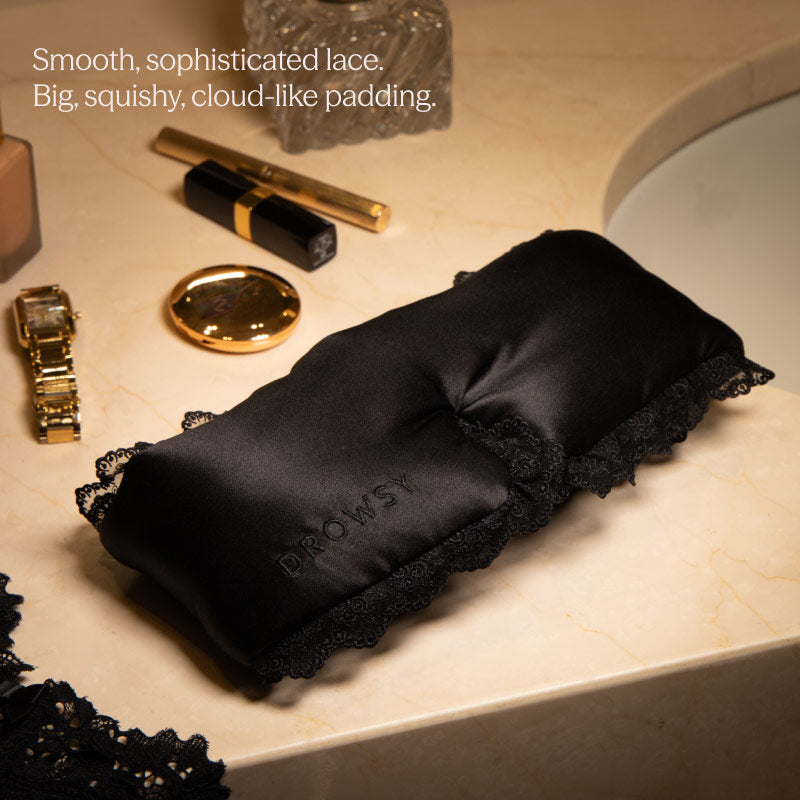 The Paris Love Story - Black Jade Sleep Mask by drowsysleepco, featuring smooth lace trim and padded, anti-aging design, rests on a marble counter near a gold watch, lipstick, compact mirror, and glass container.