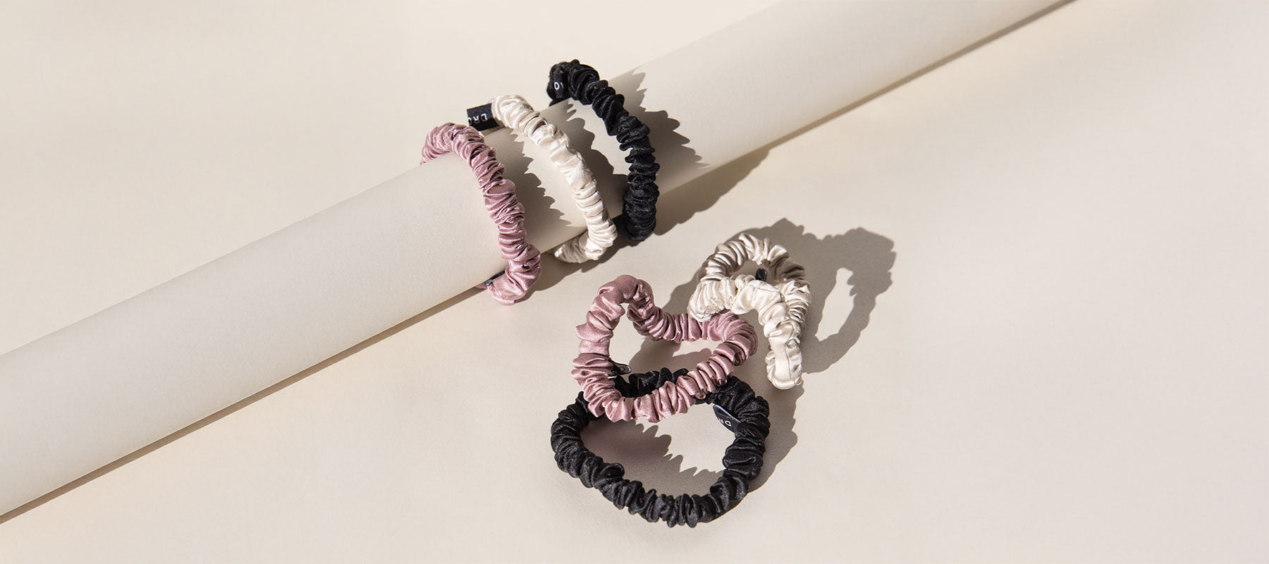 Three scrunchies in black, pink, and ivory colors are arranged on and below a cylindrical rod against a neutral beige background, creating soft shadows.
