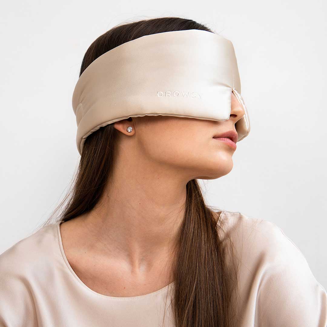 A woman with long brown hair models the drowsysleepco Sensual Sleep Bundle in Dusty Gold, featuring a beige silk sleep mask and matching satin top, posed against a plain white background.