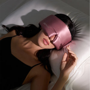 A woman in a black spaghetti strap top lies on a white pillow, wearing the drowsysleepco Damask Rose - Swarovski® embellished logo silky pink blackout eye mask. Soft light creates a serene beauty sleep vibe.