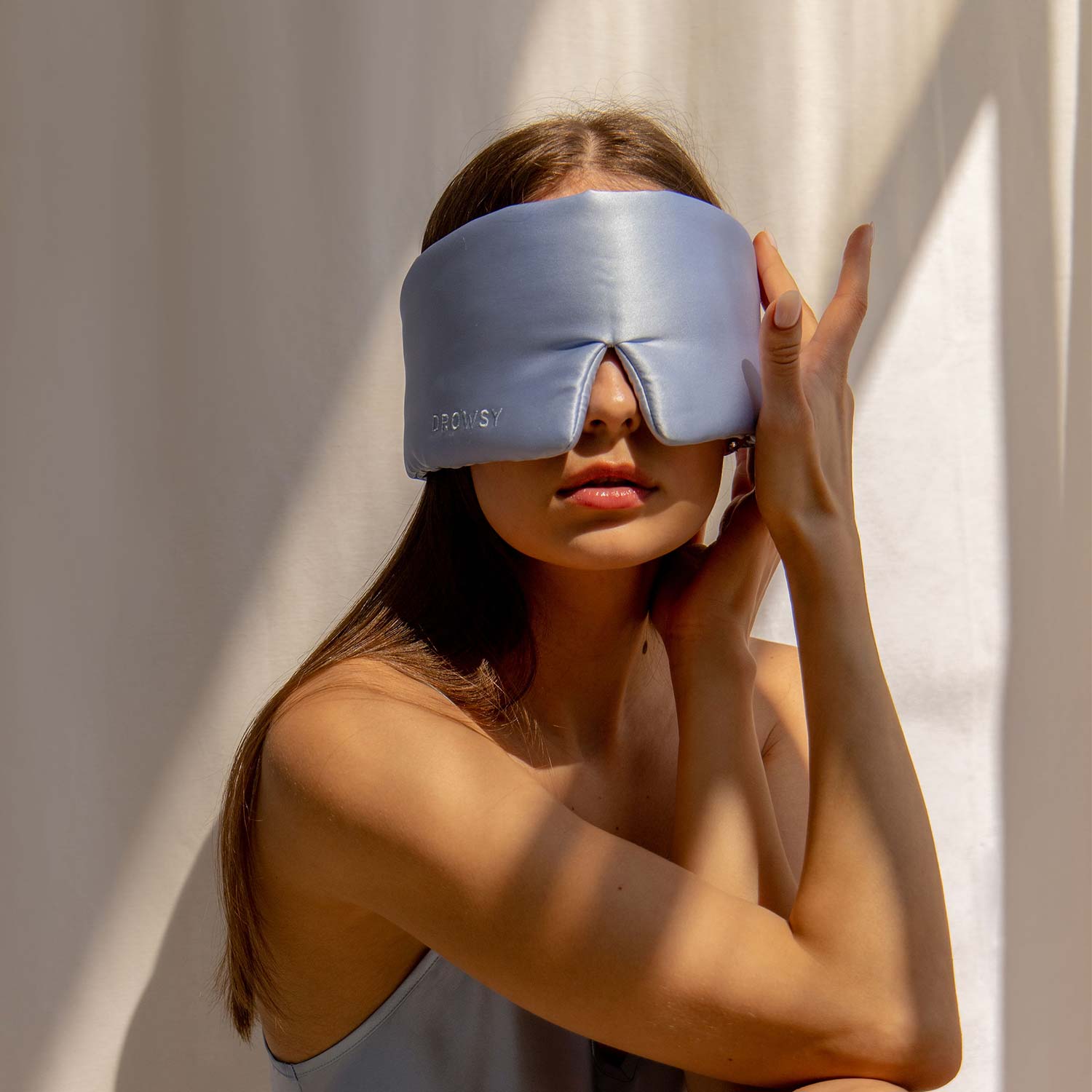 Model with baby blue coloured silk sleep mask on a white background in the sunlight