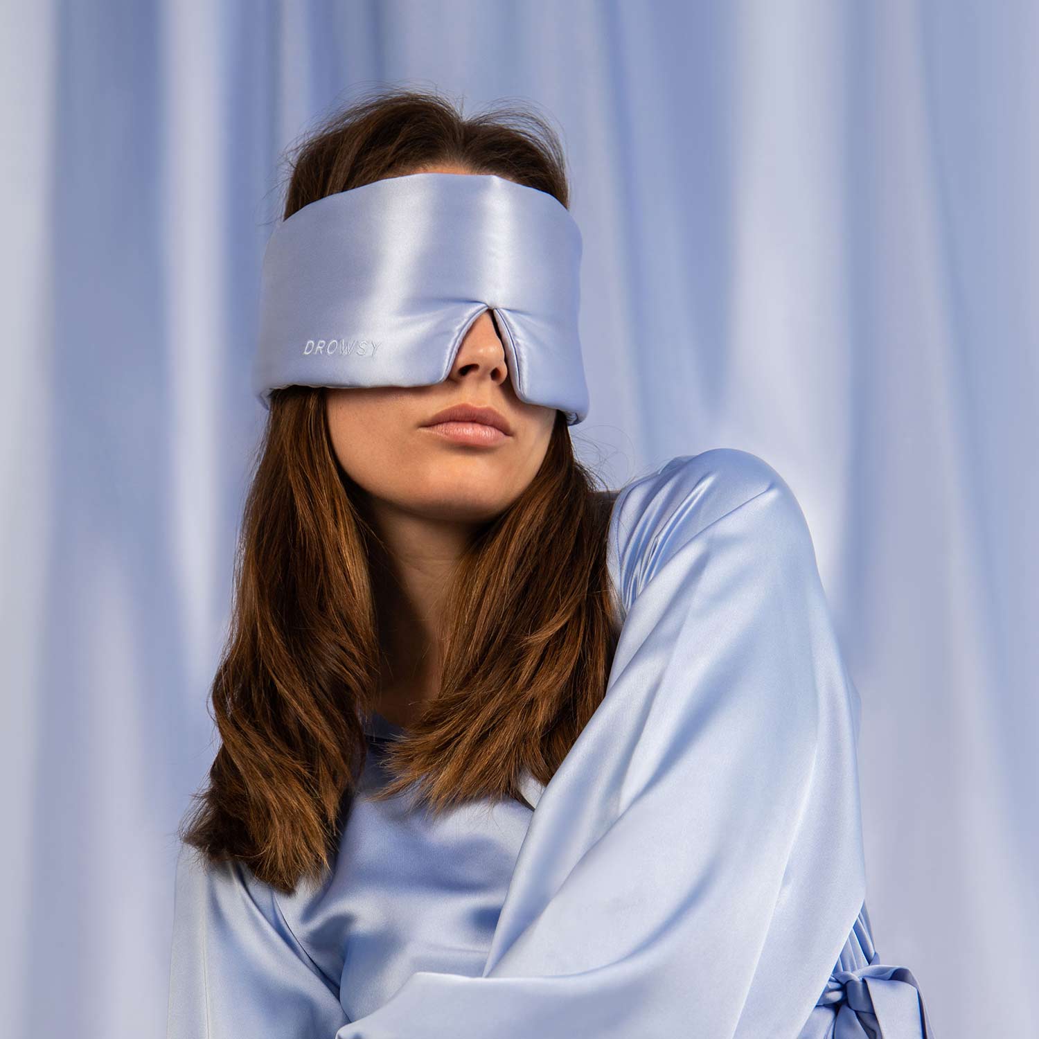 Model wearing a baby blue coloured silk sleep mask with a blue silk curtain backdrop