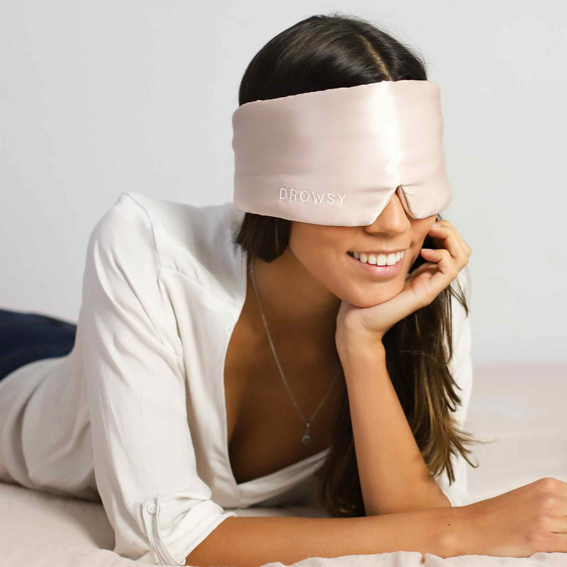 Smiling model lying on her front in bed with luxury Sunset Pink Drowsy silk sleep mask over her eyes